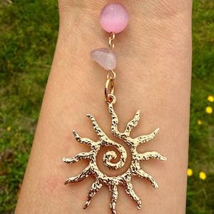 May include: A gold-colored pendant featuring a sunburst design with a spiral center. The pendant is suspended from a chain with a pink bead and a small, light-colored stone. The background is a blurred view of green grass.
