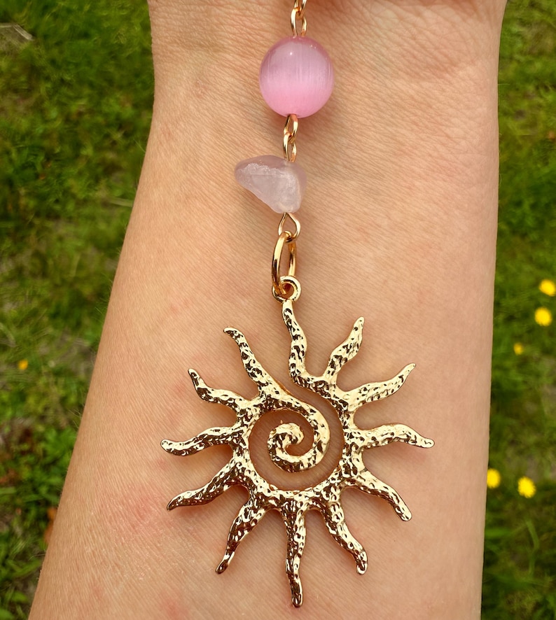 May include: A gold-colored pendant featuring a sunburst design with a spiral center. The pendant is suspended from a chain with a pink bead and a small, light-colored stone. The background is a blurred view of green grass.