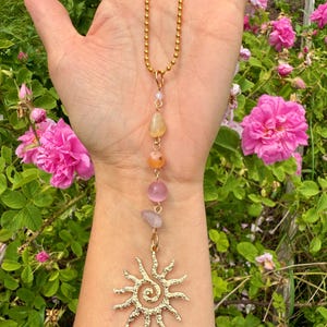 May include: Gold-colored necklace featuring a sun pendant with a spiral center and a chain of colorful gemstones. The gemstones include yellow, orange, pink, and clear stones. The necklace is displayed against a backdrop of pink roses and green foliage.