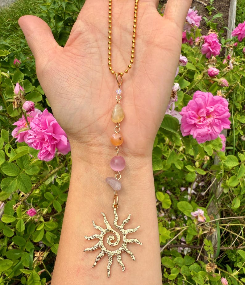 May include: Gold-colored necklace featuring a sun pendant with a spiral center and a chain of colorful gemstones. The gemstones include yellow, orange, pink, and clear stones. The necklace is displayed against a backdrop of pink roses and green foliage.