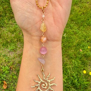 May include: A gold-colored necklace with a sun pendant and a series of colorful gemstones. The necklace features a gold chain and a pendant with a spiral design. The gemstones include yellow, orange, pink, and purple stones.