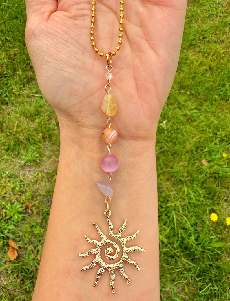May include: A gold-colored necklace with a sun pendant and a series of colorful gemstones. The necklace features a gold chain and a pendant with a spiral design. The gemstones include yellow, orange, pink, and purple stones.