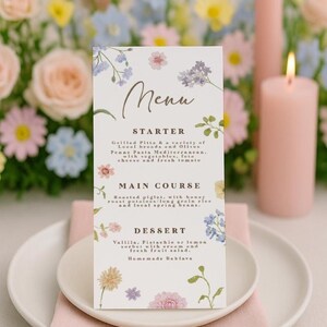 Może przedstawiać: Karta menu z motywami kwiatowymi i słowem "Menu". Menu zawiera przystawki, dania główne i desery. Karta jest ozdobiona akwarelowymi kwiatami i zielenią, umieszczona na białym talerzu z różową serwetką.
