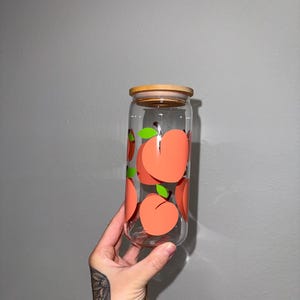 May include: A clear glass can-shaped drinking glass with a bamboo lid. The glass is decorated with repeating peach designs in shades of orange and green. The glass is held in front of a gray background.