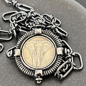 May include: A silver chain necklace with a round pendant featuring an embossed elephant design. The pendant has a textured, ornate border.