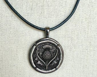 Op de afbeelding: Een zilverkleurige hangerketting met een gedetailleerd distelontwerp in een cirkelvormig frame. De hanger hangt aan een donkergroen leren koord. De distel is een symbool van Schotland en de ketting is een sieraad.