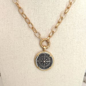 May include: A gold-toned necklace featuring a thick oval link chain and a round pendant. The pendant has a silver-toned center with a detailed compass rose design, marked with the letters N, S, E, and W, framed by a smooth gold-toned border.