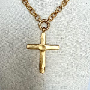 May include: Gold-tone cross pendant necklace. The cross has a textured, hammered appearance and is suspended from a chunky chain. The necklace is displayed on a neutral background.