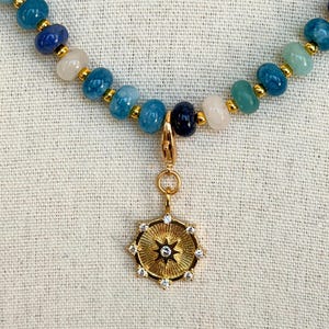 May include: A beaded necklace with a gold pendant. The necklace features blue, white, and gold beads. The gold pendant is circular with a starburst design and small clear stones. The necklace is displayed on a light-colored fabric.