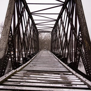 Puede incluir: Un puente metálico envejecido, de color marrón oscuro, cruza un río bajo un cielo nublado. Las tablas de madera del puente están ligeramente cubiertas de nieve, guiando la vista hacia los árboles distantes.