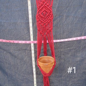 Puede incluir: Un colgador de plantas de macramé hecho a mano en color rojo intenso, diseñado para sostener una pequeña maceta de terracota. El colgador está suspendido de una varilla de bambú y presenta intrincados patrones de nudos. La maceta tiene unos 10 cm de diámetro.