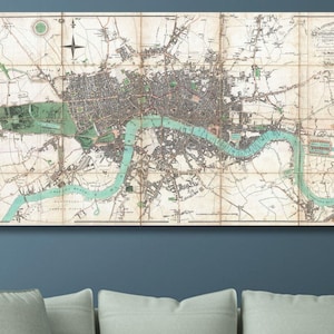 May include: An antique map of London, framed and displayed on a blue wall above a gray sofa. The map features a detailed layout of the city with a turquoise river running through it. The map is in a rectangular shape.