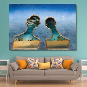 May include: A rectangular wall art print featuring two golden-framed silhouettes of human heads. Inside the frames, a surreal desert landscape with a blue sky, clouds, and small figures is visible. The artwork is displayed above a grey sofa.