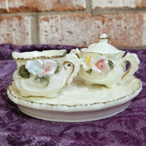 May include: A white porcelain creamer and sugar set with floral accents. The set features a delicate floral design with pink and white flowers and green leaves. The set is on a white saucer with a gold trim.