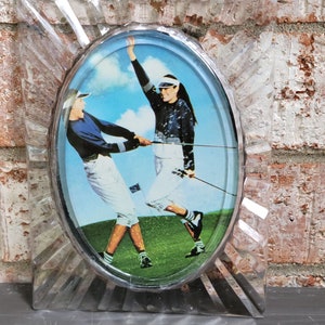 May include: A clear plastic oval frame with a picture of two people playing golf. The frame has a starburst design around the edge.