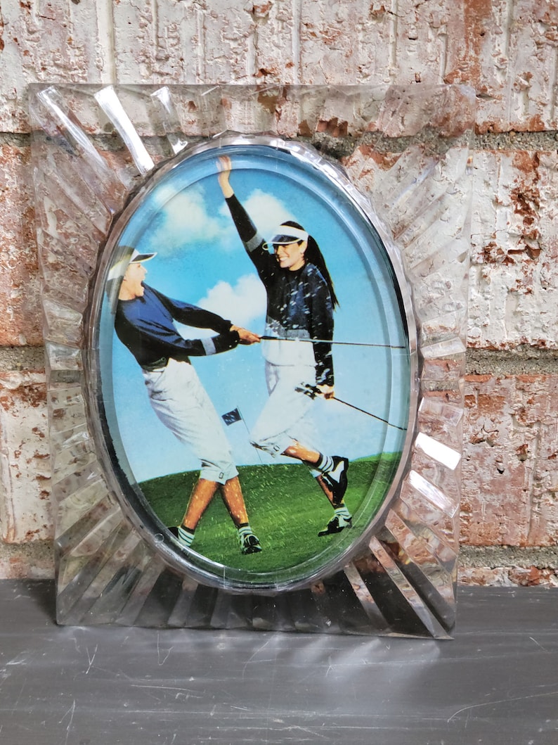 May include: A clear plastic oval frame with a picture of two people playing golf. The frame has a starburst design around the edge.