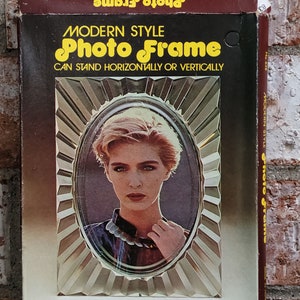 May include: A brown cardboard box with a gold and white photo frame inside. The frame is oval shaped and has a decorative design. The box has the text "Modern Style Photo Frame" and "Can stand horizontally or vertically". The box also has the dimensions "9cm x 13cm".