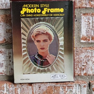 May include: A brown cardboard box with a gold and white photo frame inside. The frame is a modern style and can stand horizontally or vertically. The frame is 9 cm x 13 cm.