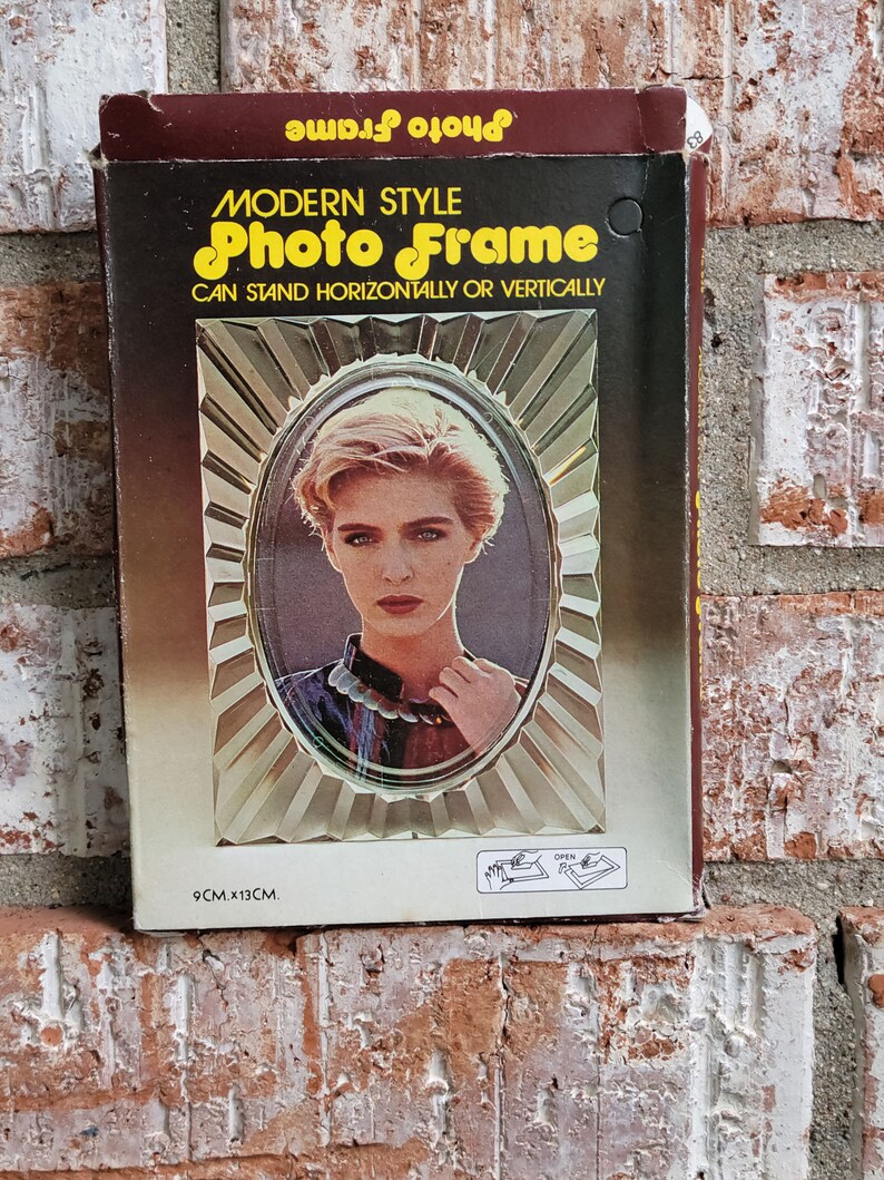May include: A brown cardboard box with a gold and white photo frame inside. The frame is a modern style and can stand horizontally or vertically. The frame is 9 cm x 13 cm.