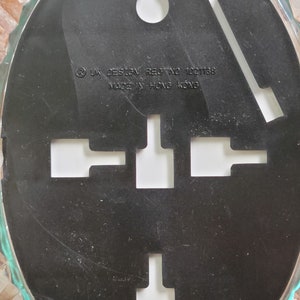 May include: Black oval-shaped wall hanging with a textured surface. It has a hole at the top for hanging and four cut-out shapes in the center. The text on the back reads "UK DESIGN REG NO 1001138 MADE IN HONG KONG".