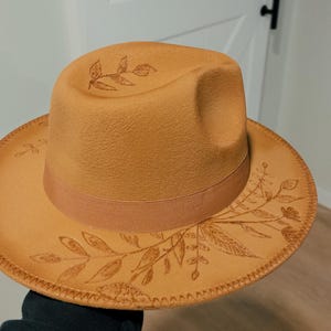 May include: A brown felt fedora hat with a brown ribbon and a floral design. The hat has a stitched edge.