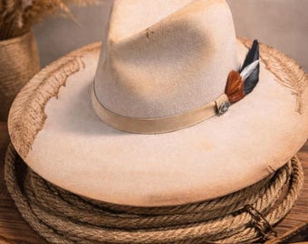 May include: A beige felt fedora hat with a wide brim featuring etched feather patterns. The hat has a light tan ribbon band adorned with a small metallic emblem and a cluster of brown, black, and white feathers on the side.