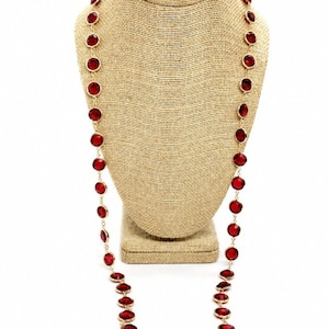 May include: A long necklace featuring a series of circular, faceted red stones set in gold-toned metal bezels. The stones are linked together in a continuous chain, creating a uniform, repeating pattern that hangs down in a long, elegant loop.
