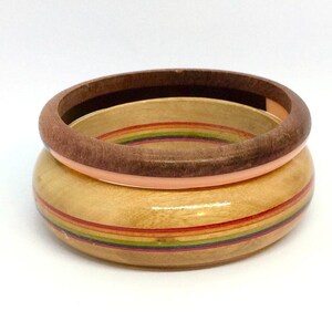 May include: Two wooden bangles. The bottom bangle is light-colored with rainbow stripes. The top bangle is dark brown. The bangles are stacked on top of each other against a white background.
