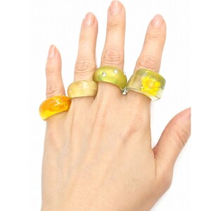 May include: Four chunky resin rings in shades of yellow and green. One ring is translucent orange-yellow, one is solid pale yellow, one is lime green with three embedded clear rhinestones, and one is clear with a yellow flower inside.