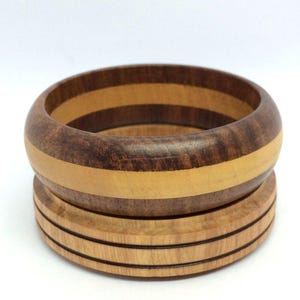 May include: A handcrafted wooden bangle bracelet with a layered design. The bracelet features alternating bands of dark and light wood, creating a striped pattern. The base has three parallel grooves. The overall design is simple and elegant.
