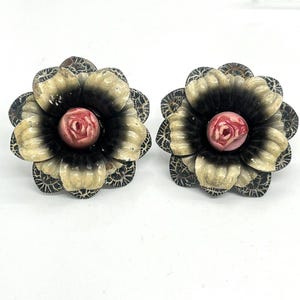 May include: A pair of flower-shaped earrings. Each earring features a black center with cream-colored petals and a pink center. The petals have a patterned edge. The earrings are set against a white background.