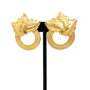May include: A pair of gold-toned earrings featuring a circular hoop base with a textured, cross-hatched pattern. Attached to the top of each hoop is a sculpted, three-dimensional leaf motif with smooth, curved edges and a polished metallic finish.