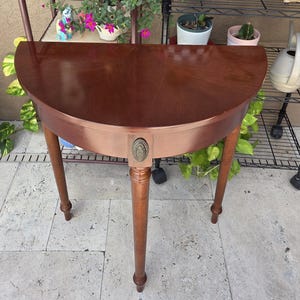 May include: A semi-circular wooden console table with a smooth, polished dark brown finish. The table features three slender, tapered legs and a single decorative oval-shaped metal medallion centered on the front apron of the furniture piece.