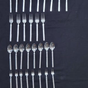 May include: A collection of stainless steel silverware, including knives, spoons, and forks, arranged on a black surface. The silverware has a simple, elegant design, suitable for formal dining. The image showcases a complete set.
