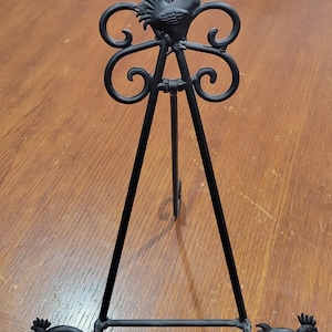 Può includere: Cavallo da cavalletto in metallo nero con un design a forma di gallo. Il cavalletto ha un design decorativo a volute e tre gambe con accenti a forma di gallo.