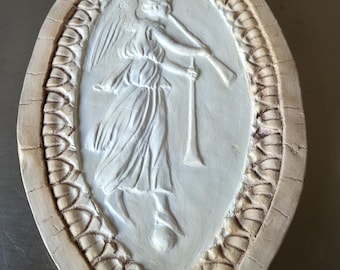 May include: Oval-shaped, off-white relief sculpture featuring an angel playing a trumpet. The angel is depicted with flowing robes and wings, set within a decorative border. The border has a cracked, aged appearance, adding to the vintage aesthetic.
