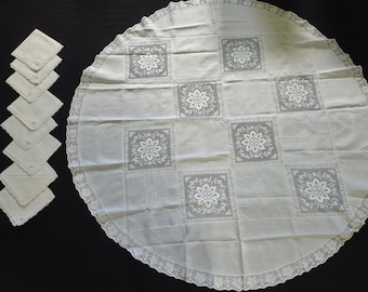 May include: A round white tablecloth featuring a repeating pattern of square panels with floral embroidery and a wide lace border, accompanied by a set of eight matching square napkins with scalloped edges and delicate corner embroidery details.