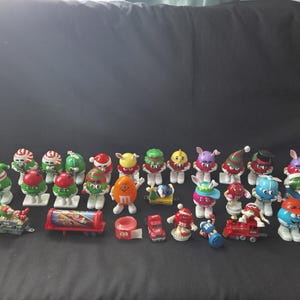 May include: A collection of colourful M&M's candy character figurines, many wearing festive hats and outfits. Some figurines are on skis, and there are also miniature vehicles like a train and a truck.