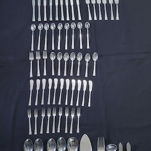 Könnte beinhalten: Ein Besteckset, bestehend aus Gabeln, Löffeln und Messern, in Reihen auf einer schwarzen Oberfläche angeordnet. Das Besteck ist aus poliertem Metall und hat ein schlichtes, elegantes Design.