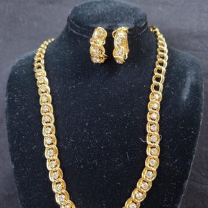 May include: A gold-toned necklace and matching hoop earrings set. The jewelry features a chain-link design with embedded clear crystals. The necklace and earrings are displayed on a black velvet bust.