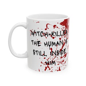 以下が含まれることがあります： 黒い文字で「WITCH-KILLER THE HUMAN IS STILL INSIDE HIM」と書かれた白いセラミックマグ。赤い飛沫のデザイン。マグには白いハンドルが付いており、温かい飲み物にも冷たい飲み物にも適しています。