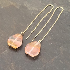 May include: A pair of gold-colored threader earrings. Each earring features a faceted, teardrop-shaped, pink and orange gemstone. The earrings are displayed on a dark gray surface.