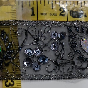 May include: Decorative trim with a gray background, featuring intricate black embroidery, sequins, and iridescent accents. The trim is approximately 1 inch wide, with a floral and leaf pattern. A yellow measuring tape is in the background.