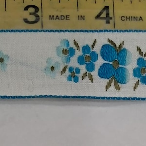 May include: White ribbon with a repeating floral pattern in shades of blue and teal, accented with brown leaves. The ribbon is bordered with a teal stitched edge. A measuring tape is in the background.