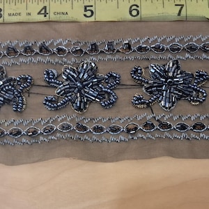 May include: A decorative trim featuring a series of beaded floral designs in shades of silver and gray. The trim is approximately 2 inches wide and is displayed against a tan fabric. The trim is measured with a yellow measuring tape.