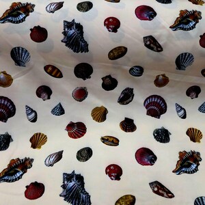 May include: A light cream-colored fabric featuring a repeating pattern of various seashells in shades of brown, red, black, and white. The print includes diverse shell shapes such as conch, scallop, and spiral varieties scattered across the surface.