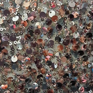 May include: Close-up of a fabric covered in a dense pattern of round, metallic sequins. The sequins vary in color, including silver, bronze, and copper, creating a shimmering effect. The sequins are attached to a fine, mesh backing.