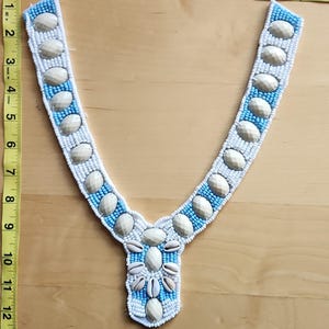 May include: A beaded collar with a V-neck design. The collar features white, light blue, and beige beads and shell accents. The collar is approximately 10 inches long.