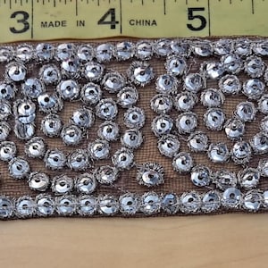 May include: A decorative trim featuring a repeating pattern of silver-colored, circular embellishments. The trim is approximately 1 inch wide and is set against a sheer, tan-colored backing. The embellishments are evenly spaced.