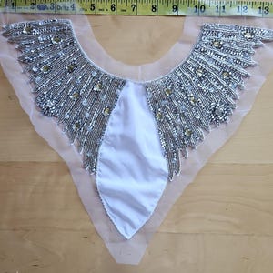 May include: A silver beaded collar with a white satin center and sheer backing. The collar is approximately 13 inches wide and features intricate beadwork in a fan-like design, with a mix of silver and gold beads.